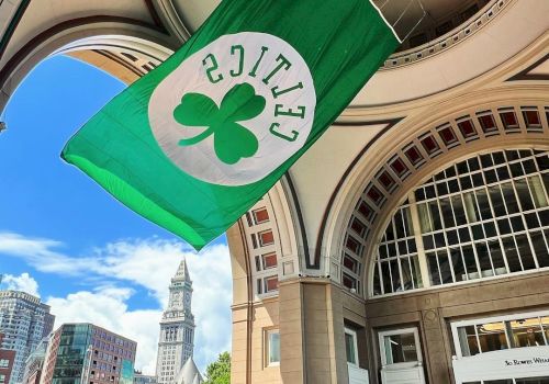 The image shows a green Celtics flag with a shamrock hanging under an arched structure, with a cityscape and clock tower in the background.