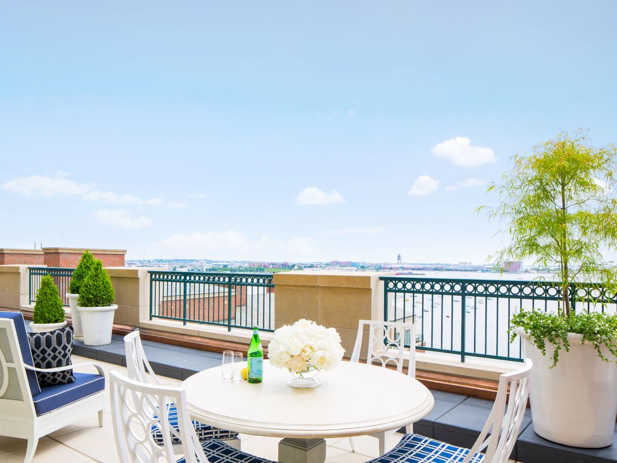 A rooftop terrace with white furniture, a potted plant, decorative cushions, and a round table with flowers and a green bottle, overlooking a scenic view.