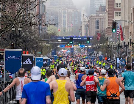 A crowded road race with many runners in colorful outfits, starting/finishing barriers, and city buildings in the background, cheering spectators, and race banners.