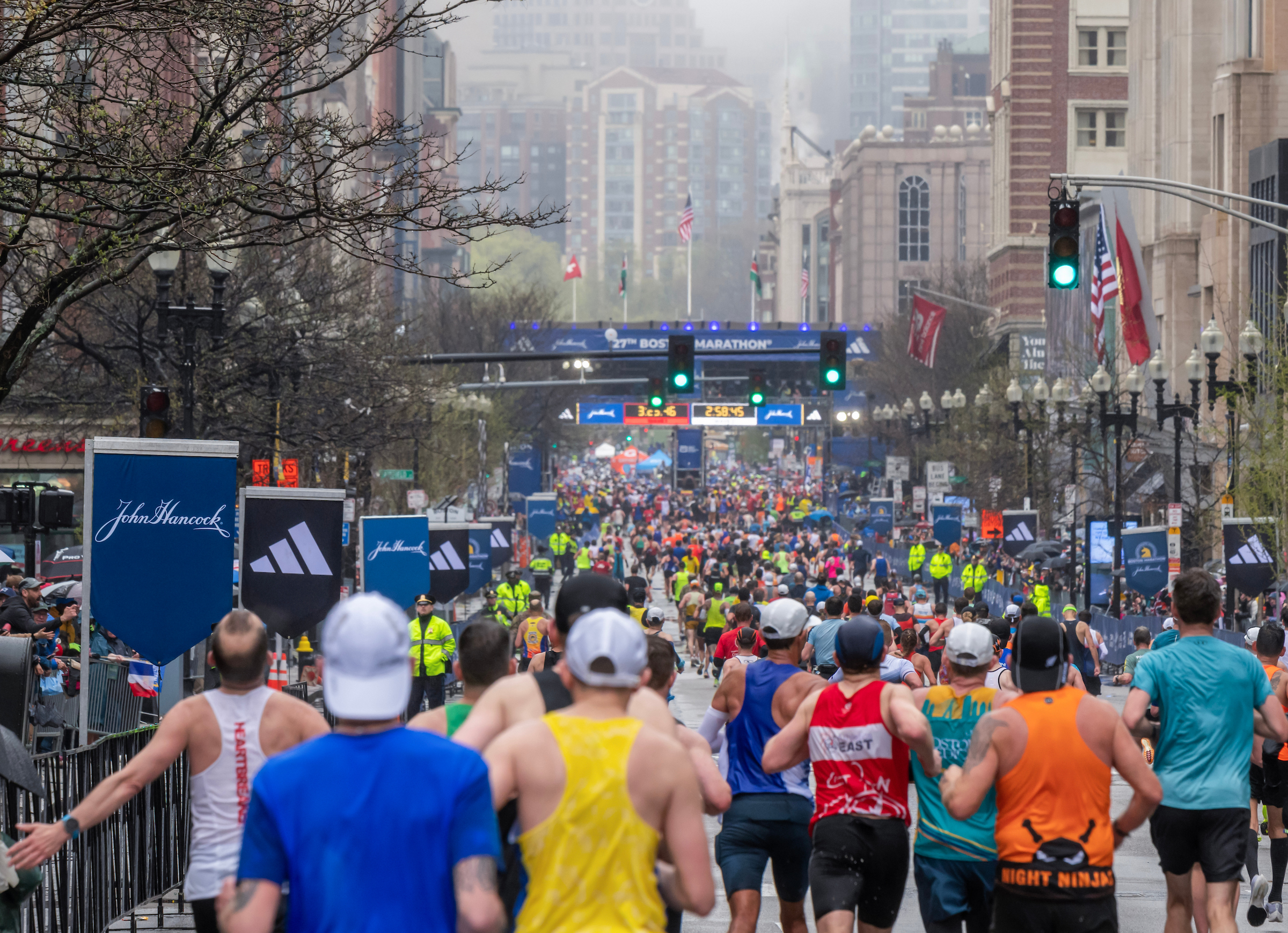 A crowded road race with many runners in colorful outfits, starting/finishing barriers, and city buildings in the background, cheering spectators, and race banners.