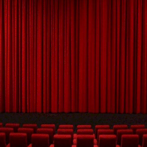 It shows a theater stage with closed red curtains and rows of red seats in the foreground.