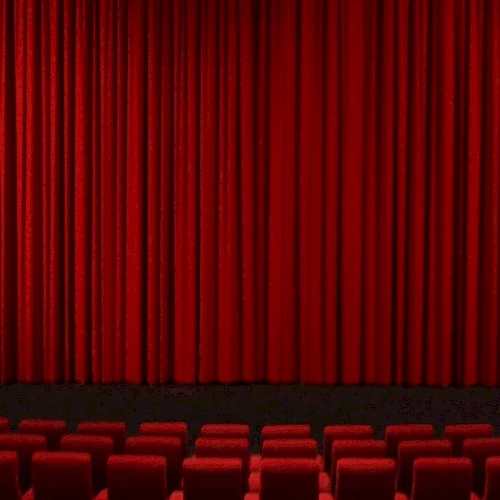 It shows a theater stage with closed red curtains and rows of red seats in the foreground.