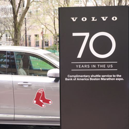 A silver car with a Boston Red Sox logo sits next to a tall sign that says &ldquo;VOLVO 70 YEARS IN THE US&rdquo; near a tree-lined street.