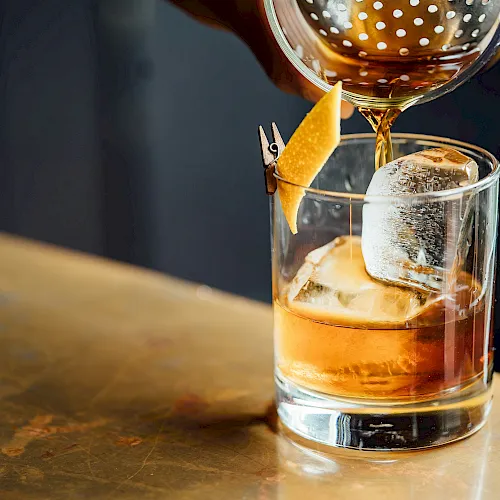 A bartender pours a golden amber drink from a cocktail shaker into an ice-filled glass, garnished with a lemon slice.