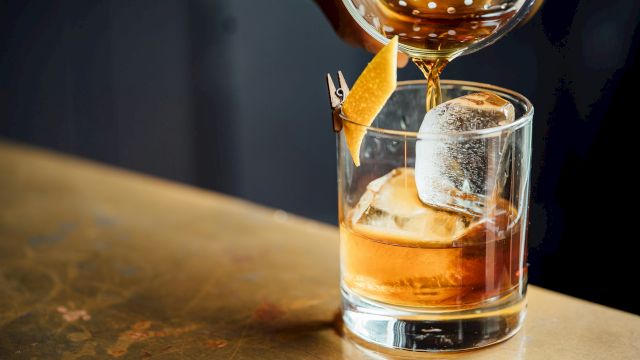 A bartender pours a golden amber drink from a cocktail shaker into an ice-filled glass, garnished with a lemon slice.
