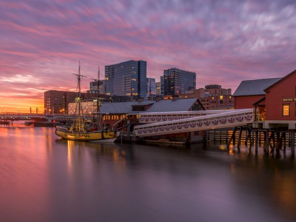 A sunset over a harbor with calm water, modern buildings in the background, a docked vintage boat, and a red wooden pier structure.