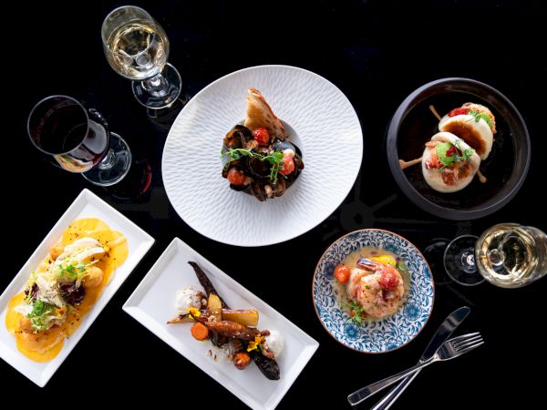 An elegant spread featuring gourmet dishes, assorted small plates, and glasses of white wine on a black background.