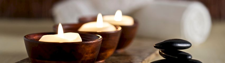 Three lit candles in wooden bowls on a rustic board, with smooth black stones stacked in the foreground, suggesting a calming balance.