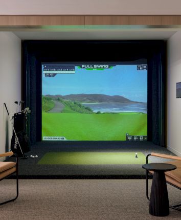 A cozy home golf simulator setup with a screen showing a sunny course, two chairs, a small table, and equipment surrounding a framed screen.