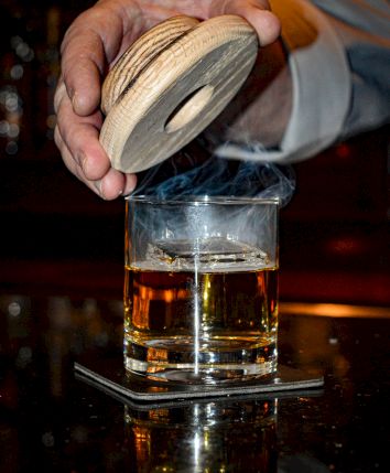 A bartender opens a wooden lid to pour whiskey into a glass at a bar, with a coaster underneath.