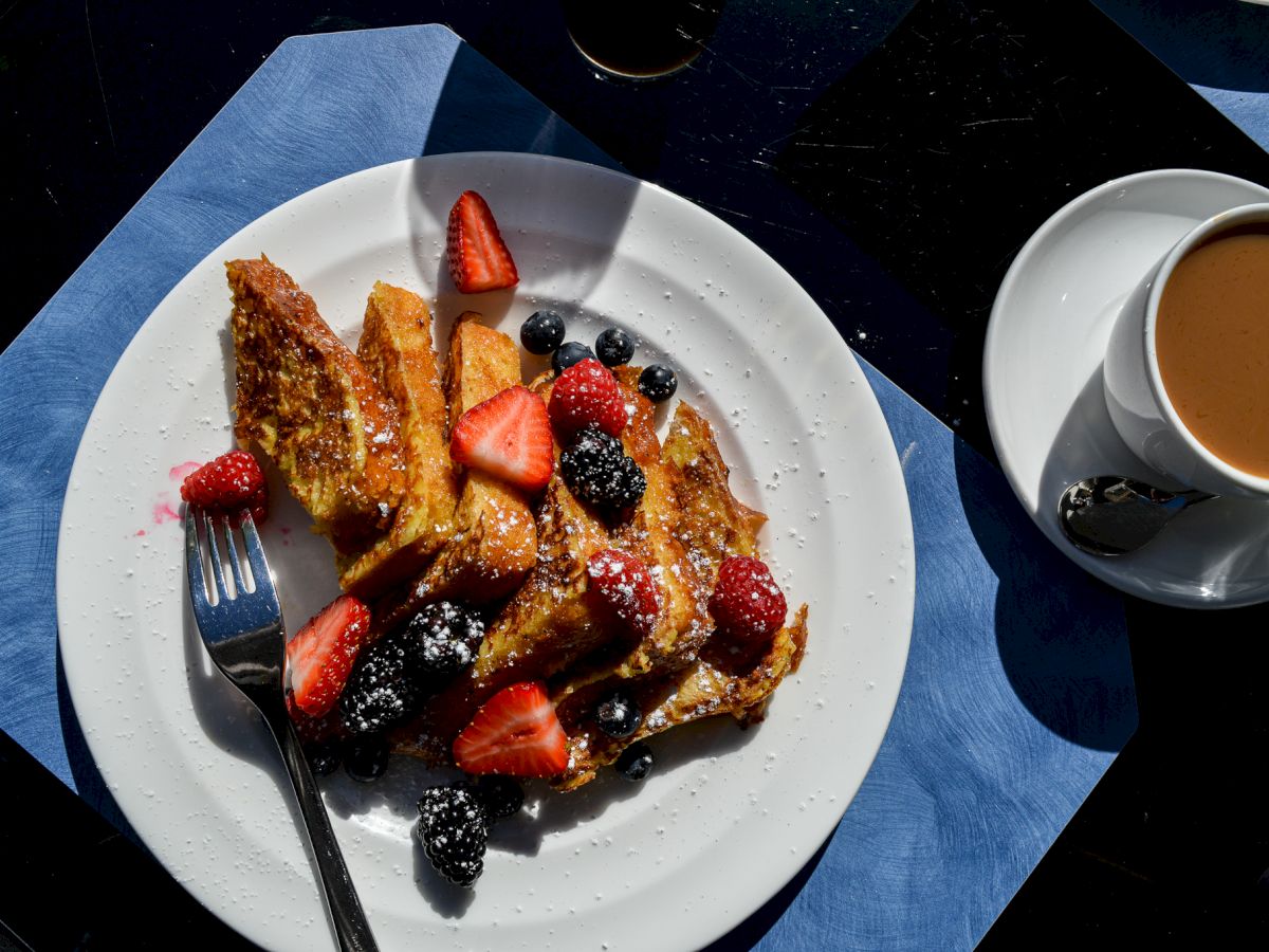 A plate of French toast topped with berries (strawberries, blueberries) and powdered sugar, with a cup of coffee on the side.