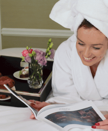 A woman in a white bathrobe and towel reading a book, with a breakfast tray of pastries and flowers on a bed.