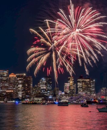 Fireworks over a city skyline at night, reflected on the water.