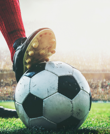 A close-up of a soccer ball on the grass with a player's boot ready to kick, stadium lights glowing in the background, action about to start.