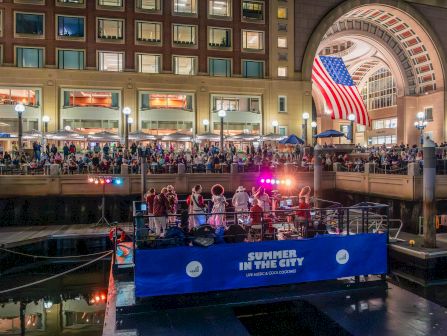 A lively waterfront scene at a city canal, with a blue party boat labeled &ldquo;Summer in the City&rdquo; docked near a grand arched bridge and a bustling crowd enjoying lights.