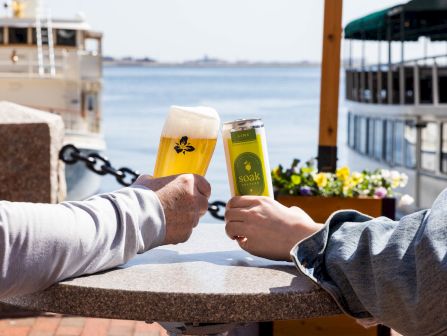 Two people clink beer glasses at an outdoor waterfront table, with boats and a calm harbor in the background.