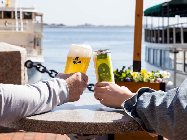Two people clink beer glasses at an outdoor waterfront table, with boats and a calm harbor in the background.