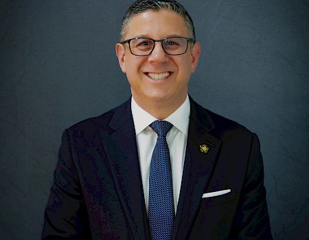 A well-dressed man in a dark suit and tie, smiling confidently in a studio portrait.