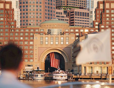 A harbor scene with a large arched gateway, tall brick buildings, a blurred foreground person, and a white sailing boat by the dock, urban waterfront.