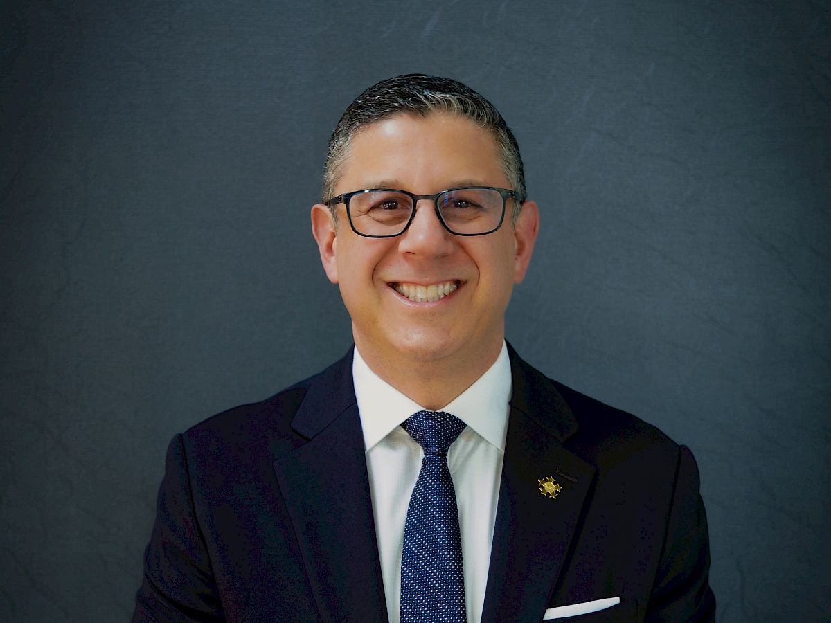 A smiling man in a dark suit and tie, with glasses, posed against a dark background, looking confident and professional.