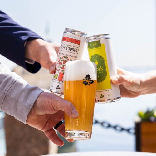 People cheers with three beer bottles and a glass of beer outdoors, celebrating a shared toast by the water.