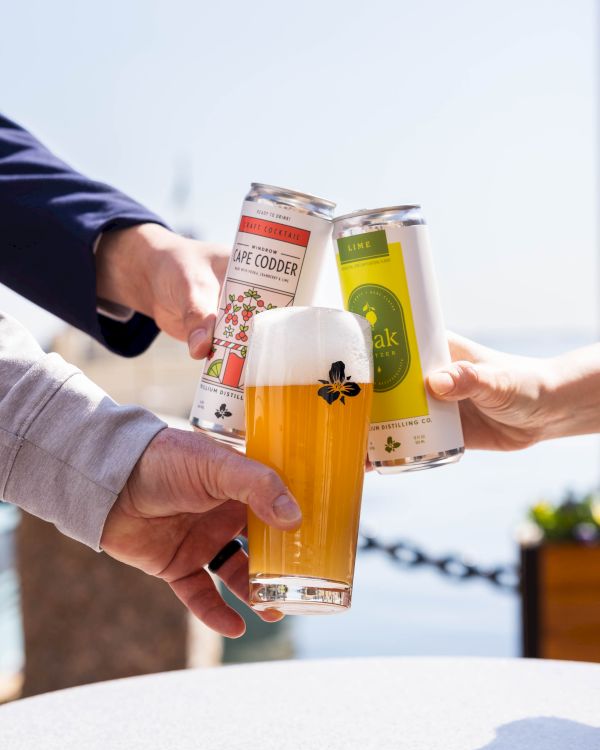 People cheers with three beer bottles and a glass of beer outdoors, celebrating a shared toast by the water.