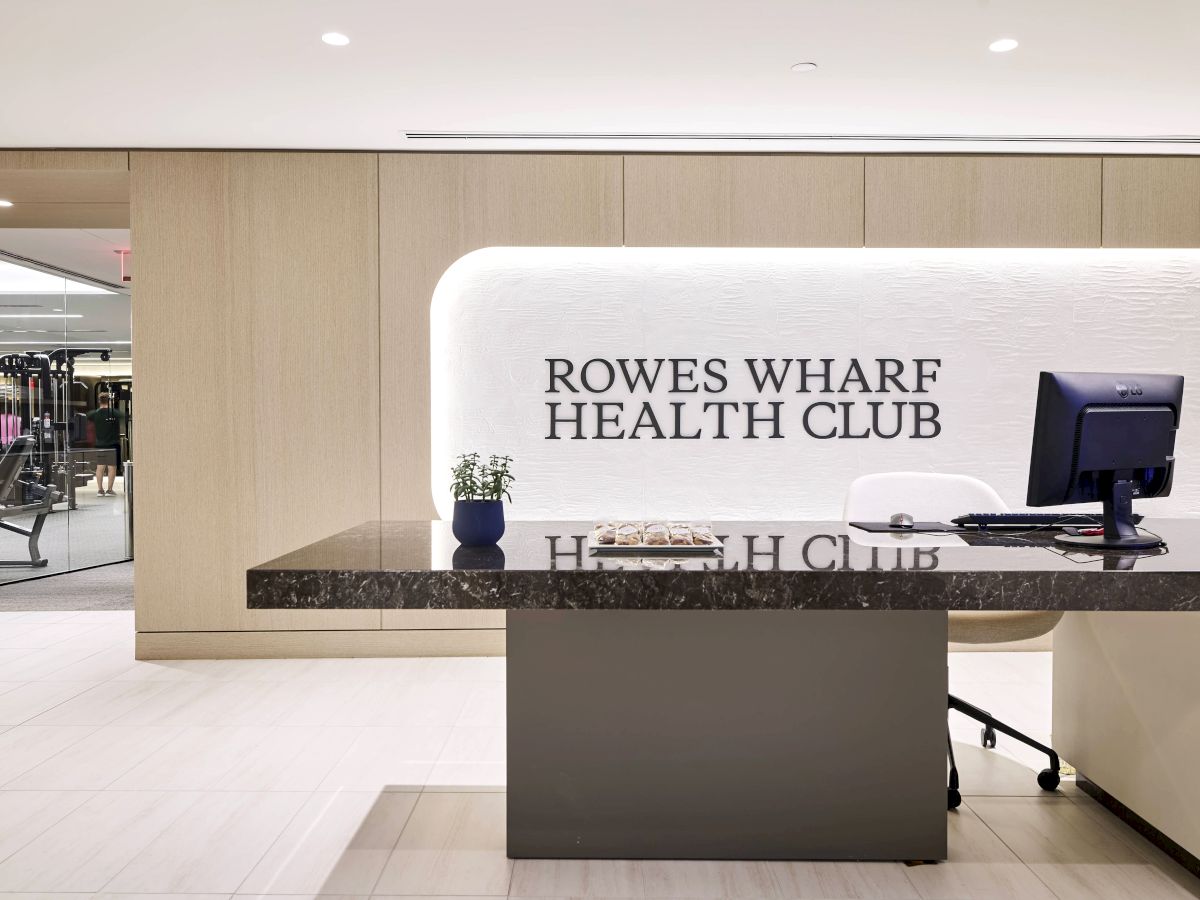 The image shows a reception desk at Rowes Wharf Health Club, featuring a computer, plant, and a modern interior design.