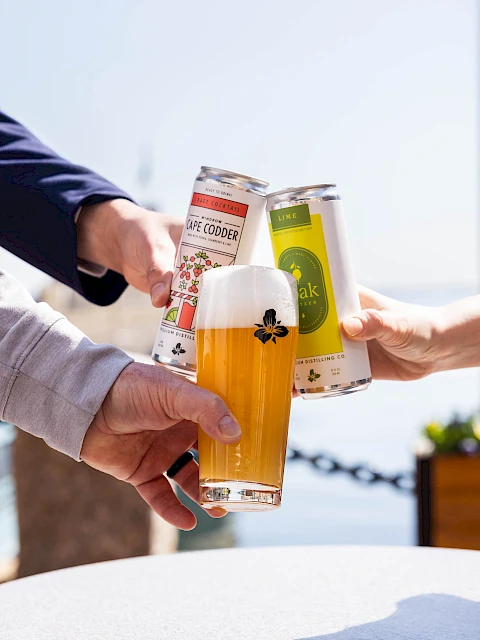 Three people cheers with cans and a glass of beer, clinking triumphantly against a sunny outdoor backdrop.