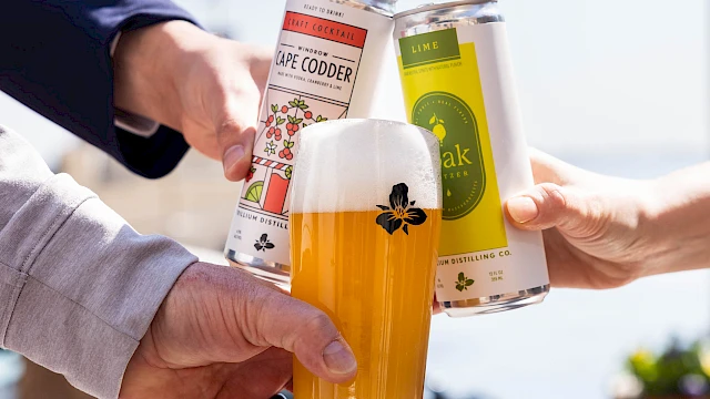 Three people cheers with cans and a glass of beer, clinking triumphantly against a sunny outdoor backdrop.