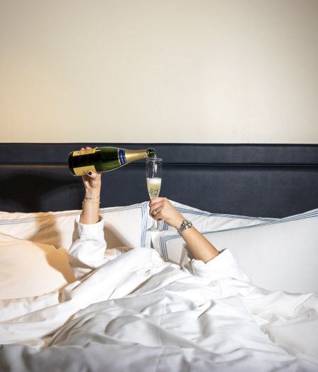 A person in bed pops champagne with a corkscrew bottle, celebrating while lying under white sheets and pillows.