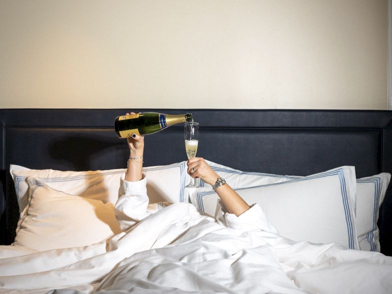 A person in bed pops champagne with a corkscrew bottle, celebrating while lying under white sheets and pillows.