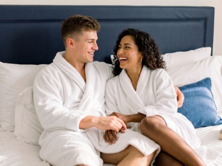 Two people in white robes sit on a bed, smiling and holding hands, cozy moment in a bright, comfortable bedroom.