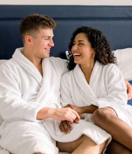 Two people in white robes sit on a bed, smiling and holding hands, cozy moment in a bright, comfortable bedroom.
