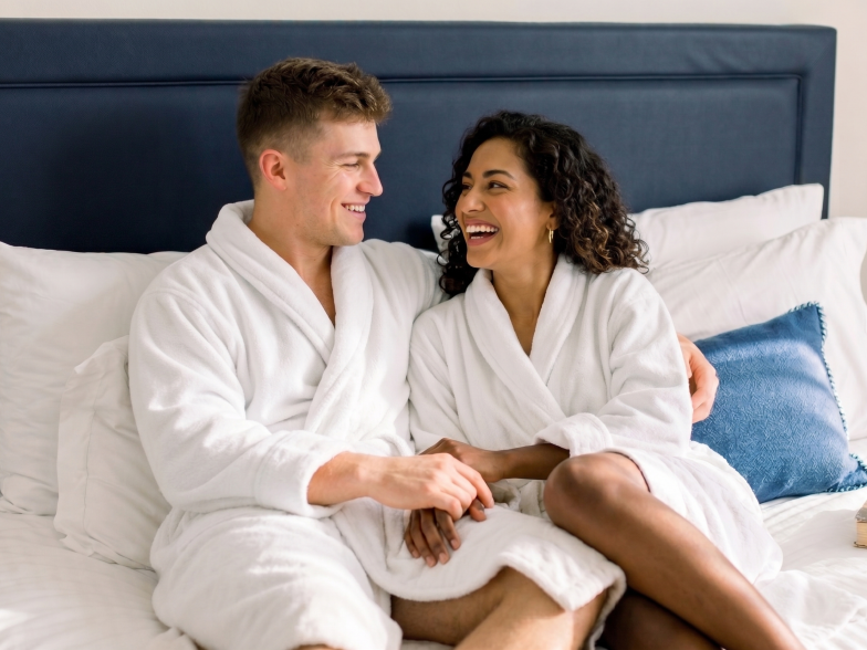 Two people in white robes sit on a bed, smiling and holding hands, cozy moment in a bright, comfortable bedroom.