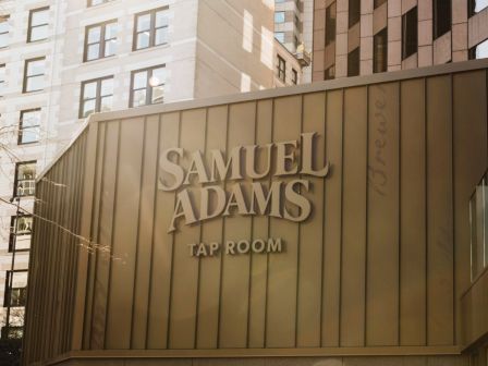 The image shows the exterior of a building with the sign "Samuel Adams Tap Room" on it, surrounded by other urban structures.