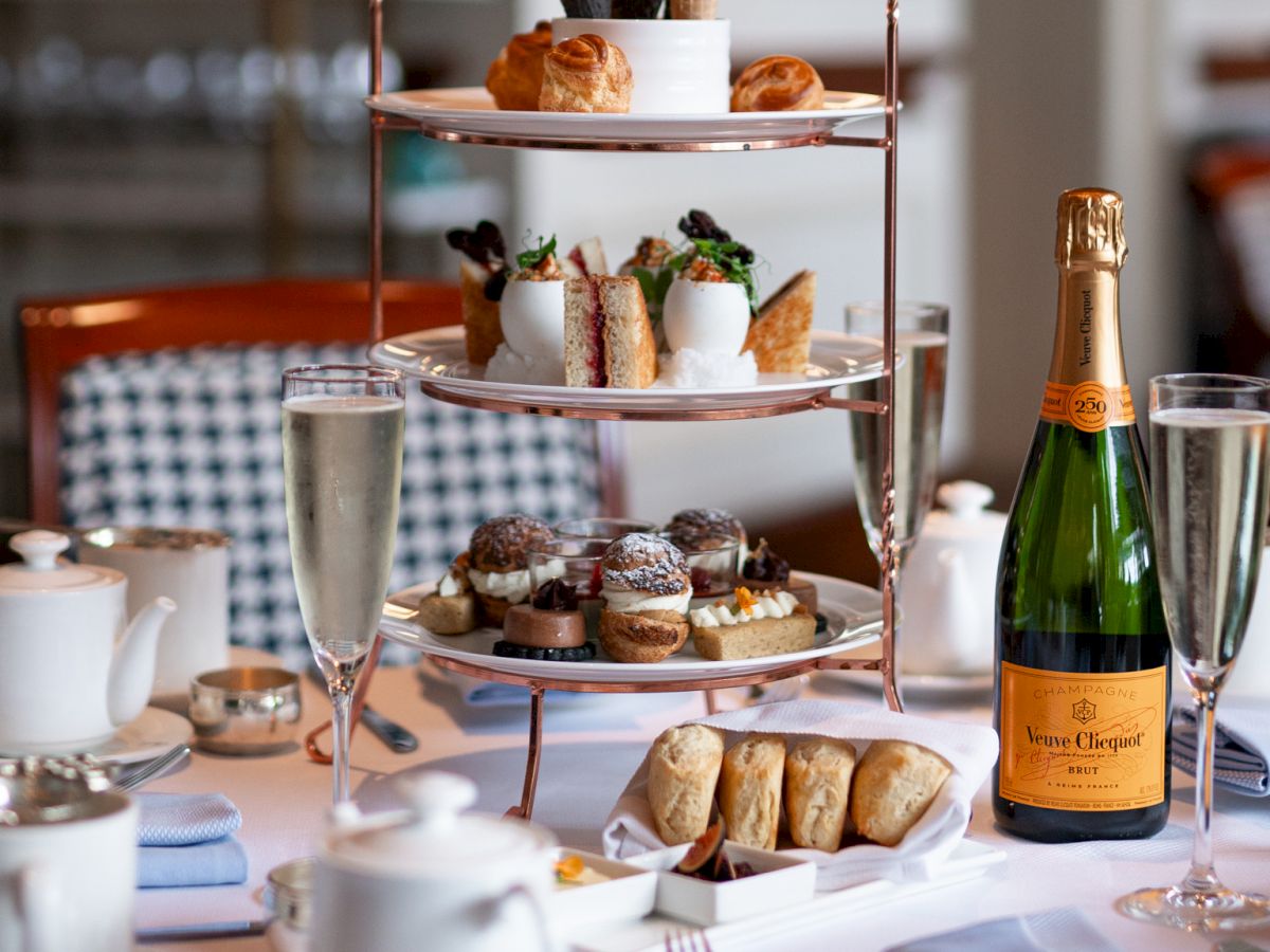 An elegant afternoon tea table setup with a tiered tray of pastries, sandwiches, a bottle of champagne, teacups, and glasses.