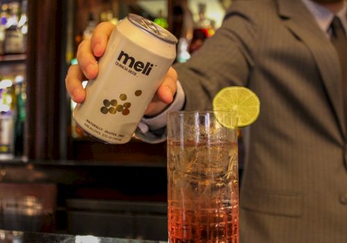 A person in a suit holding a can labeled "meli" near a drink garnished with a slice of lime in what appears to be a bar setting.