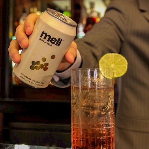 A person in a suit holding a can labeled "meli" near a drink garnished with a slice of lime in what appears to be a bar setting.