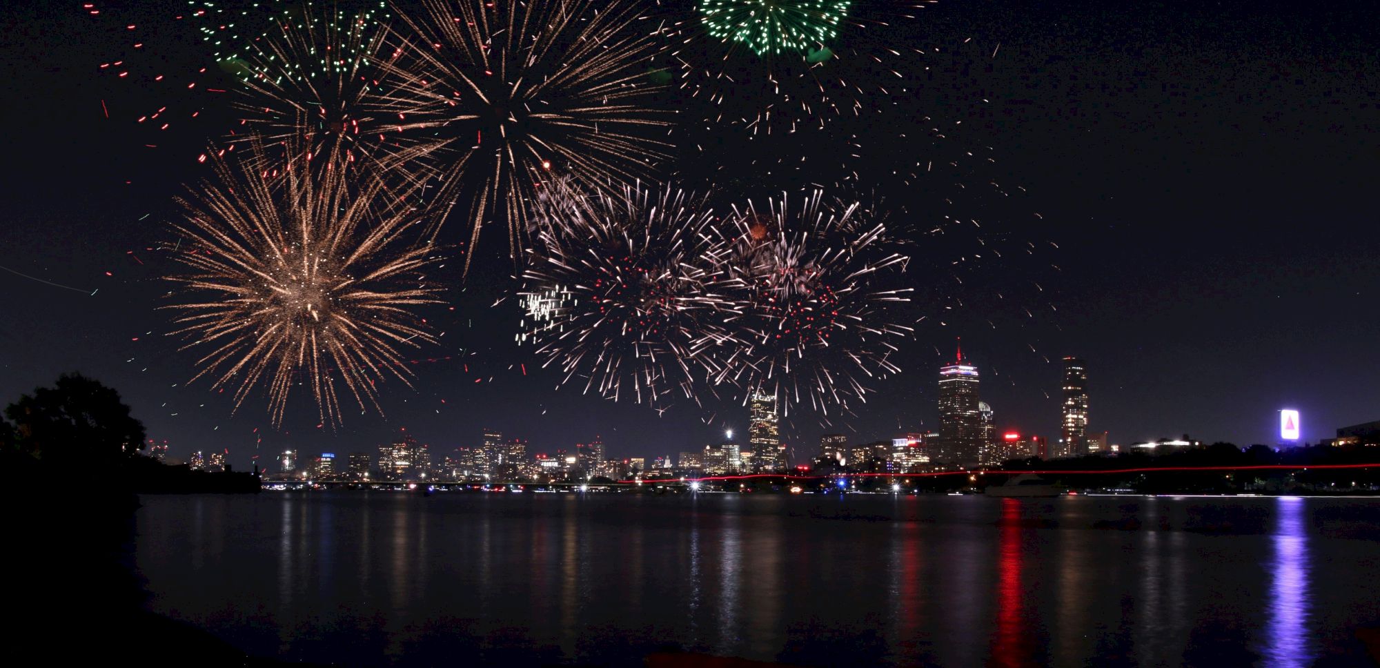 Fireworks explode over a city skyline at night, reflecting in the water.
