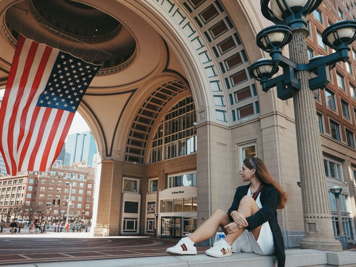 A person sits on a brick ledge under a grand arched entrance, with an American flag hanging nearby, urban buildings in the background.