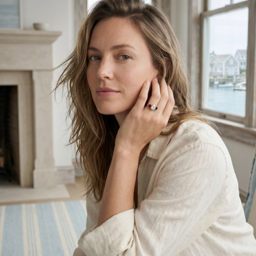 A woman with long light brown hair sits by a window in a bright room, resting her head on her hand, wearing a light-colored top.