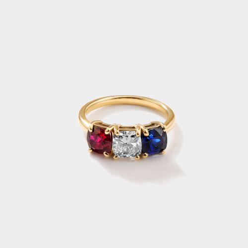 A gold ring with three gemstones: red ruby, white diamond, and blue sapphire, set in a row with a central diamond.