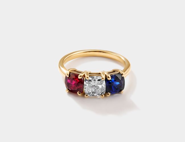 A gold ring with three gemstones: red ruby, white diamond, and blue sapphire, set in a row with a central diamond.