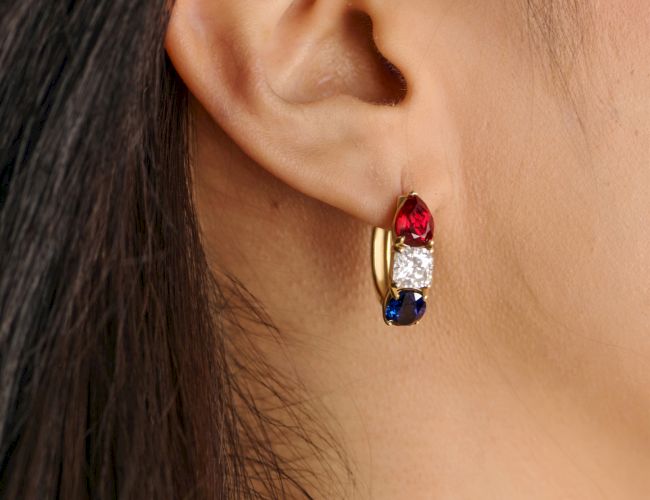 A small hoop earring with three colored stones (red, white, blue) set in a gold band, worn on the ear.