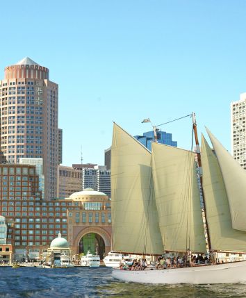 A sailboat with tall beige sails glides beside a city harbor, modern high-rises lining the waterfront under a bright blue sky.