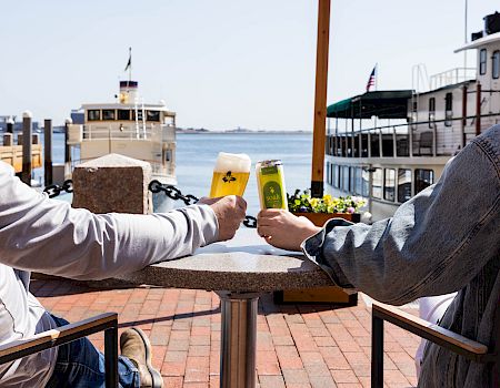Two people clink drinks at an outdoor dockside table, boats and water in the background, sunny day, casual vibe.
