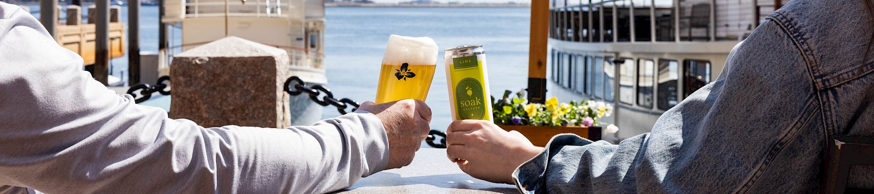 Two people toast drinks on a sunny pier, with boats and water in the background as they sit at a round table.