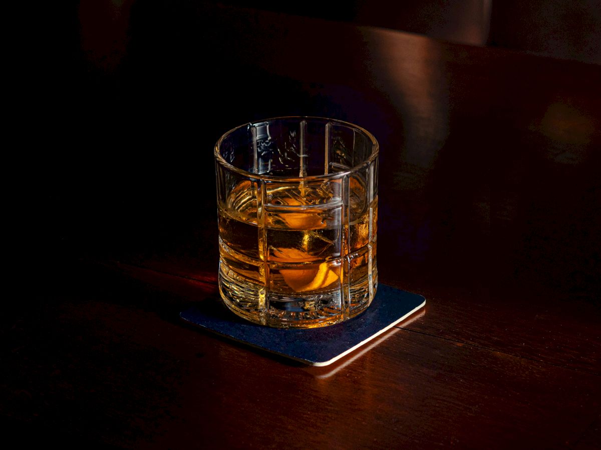 A glass of whiskey on a dark bar, resting on a blue coaster. The dim lighting highlights the amber liquid and short, chunky tumbler.