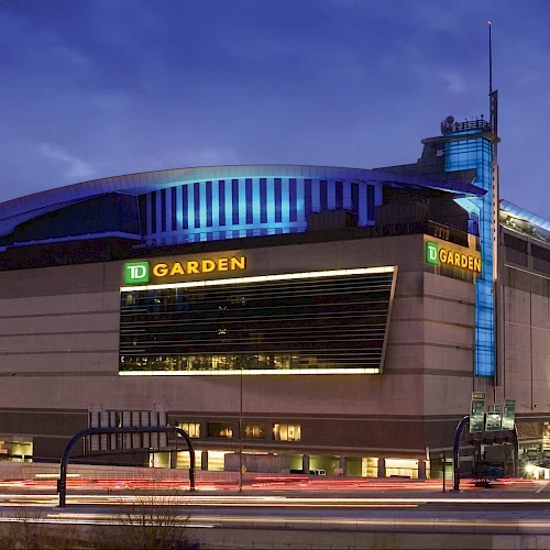 An illuminated arena with "TD Garden" signage, surrounded by city buildings with light trails from moving cars, depicted during twilight.