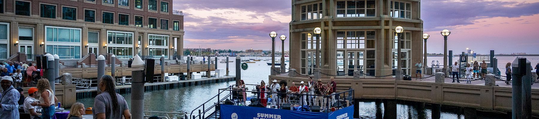 People sit along a waterfront dining area near a domed pavilion by the pier, watching a blue boat perform on the water at dusk.